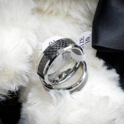 Two rings with 3K carbon fiber detailing and shiny metallic bands, each attached to a white size tag marked "SIZE 10", resting on a soft, fluffy white surface with black fabric visible nearby.