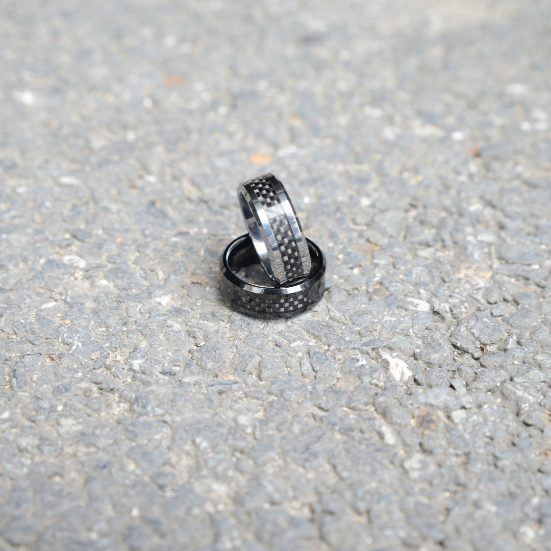 Two rings with 3K carbon fiber detailing and shiny metallic accents, stacked on a rough gravel surface.
