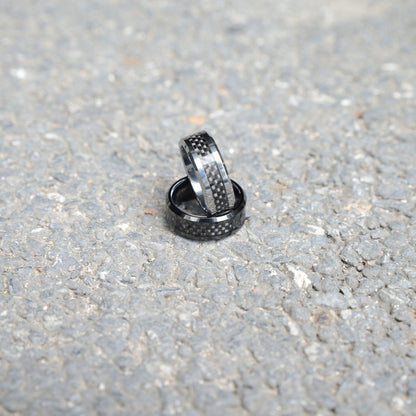 Two rings with 3K carbon fiber detailing and shiny metallic accents, stacked on a rough gravel surface.