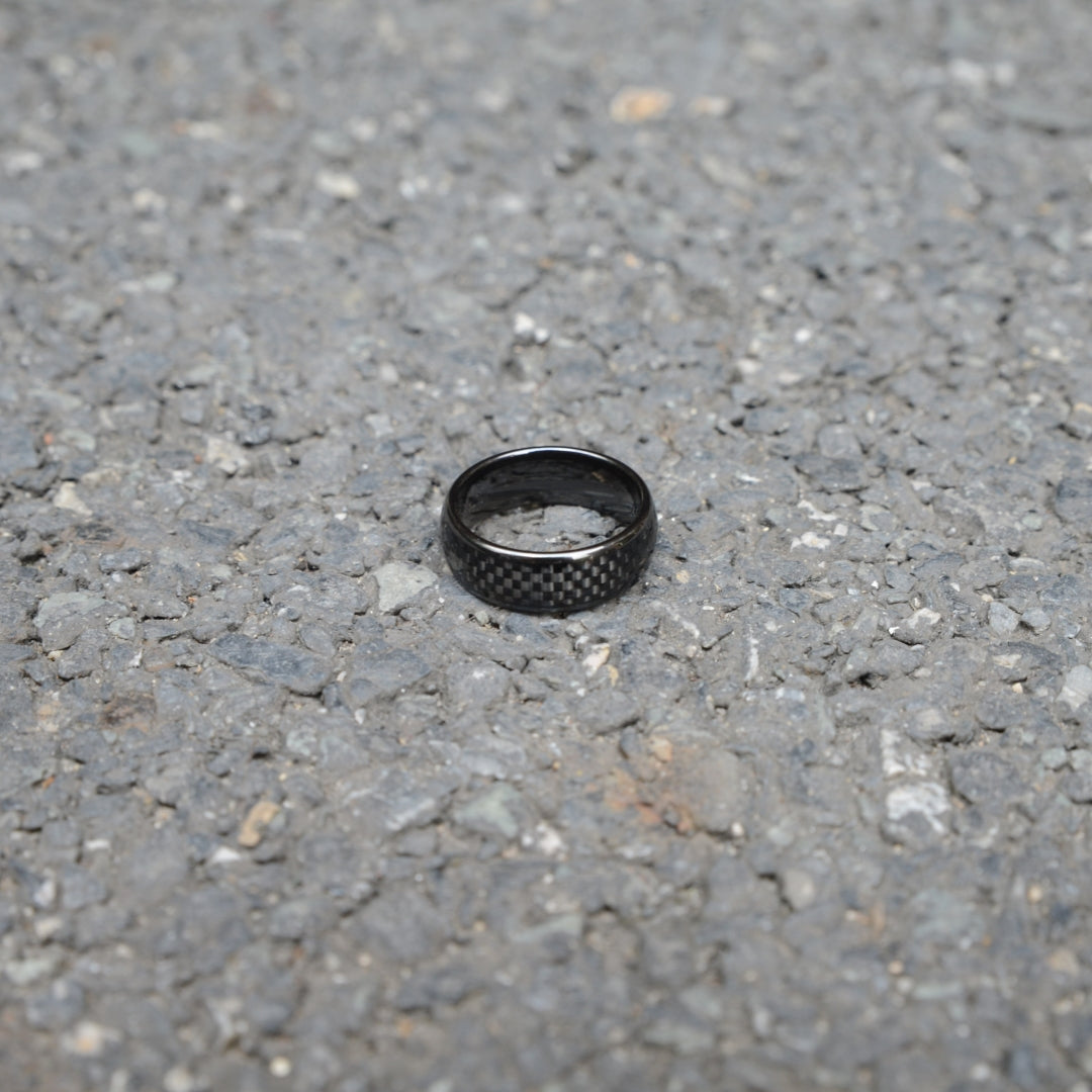 A black ring featuring 3K carbon fiber detailing, resting on a rough gravel surface.