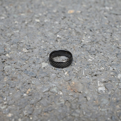 A black ring featuring 3K carbon fiber detailing, resting on a rough gravel surface.