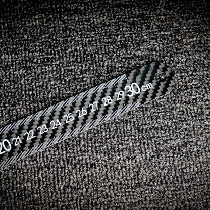 Close-up of a 3K carbon fiber ruler with clear measurement markings up to 30 cm, placed on a textured surface.