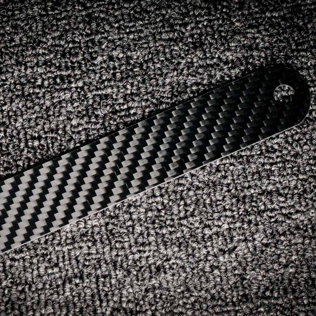 Close-up of the back side of a 3K carbon fiber ruler with a distinct weave pattern, placed on a textured surface.