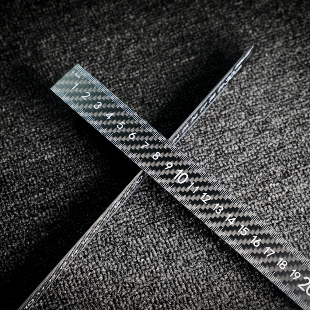 Two 3K carbon fiber rulers with distinct weave patterns and clear measurement markings, crossed on a textured dark surface.