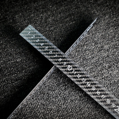 Two 3K carbon fiber rulers with distinct weave patterns and clear measurement markings, crossed on a textured dark surface.