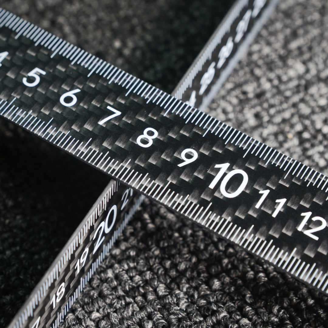Close-up of two 3K carbon fiber rulers with distinct weave patterns and clear measurement markings, crossed over each other on a textured surface.