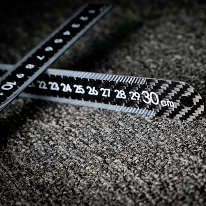 Close-up of two 3K carbon fiber rulers with distinct weave patterns and clear measurement markings (up to 30 cm visible), arranged crosswise on a textured dark surface.