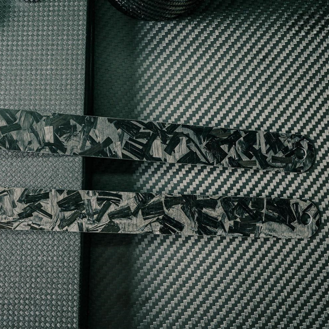 Close-up of the back sides of two forged carbon fiber rulers with distinct patterns, placed on a carbon fiber textured surface.