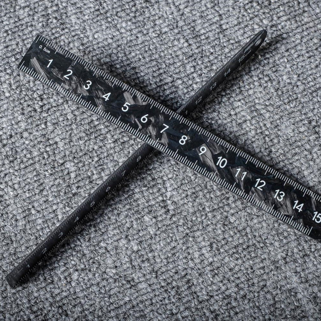 Close-up of two forged carbon fiber rulers with distinct patterns and clear measurement markings, crossed on a textured surface.