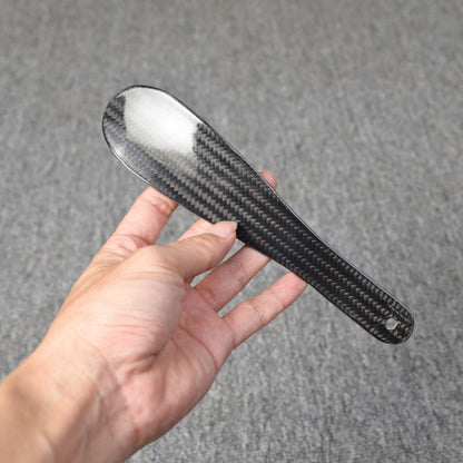 Close-up detail of a short 3K carbon fiber shoe horn with a distinct woven pattern, held in a hand against a textured gray background.