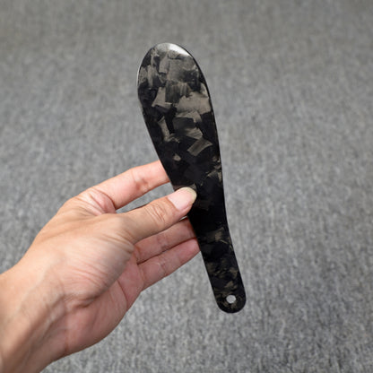 Close-up detail of a short forged carbon fiber shoe horn with a distinct chopped texture, held in a hand against a gray textured background.