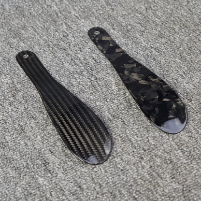Close-up detail of two short carbon fiber shoe horns—one with a 3K carbon fiber woven pattern and the other with a forged carbon fiber chopped texture—placed on a gray textured surface.