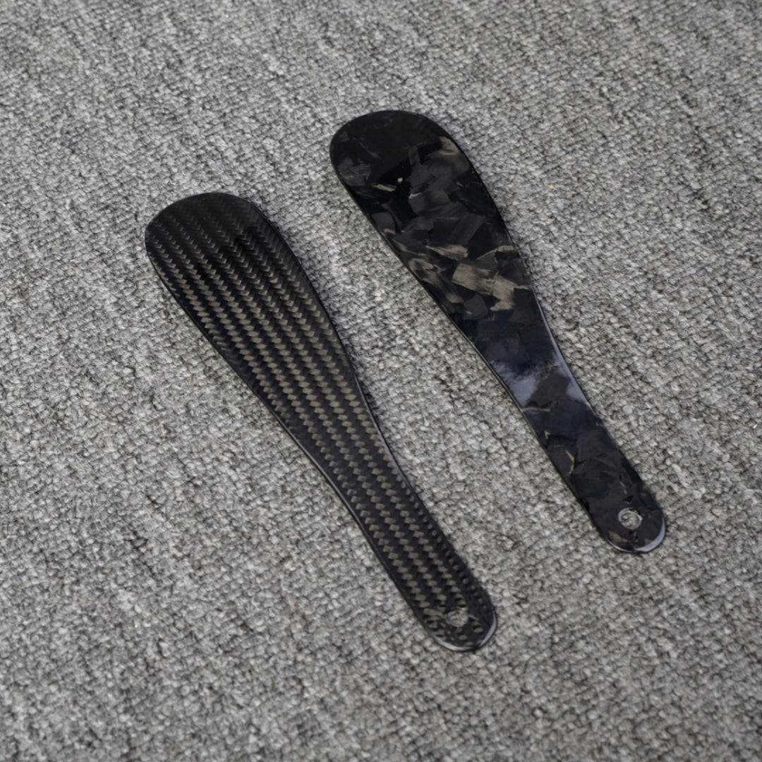 Close-up detail of two short carbon fiber shoe horns—one featuring a 3K carbon fiber woven pattern and the other showcasing a forged carbon fiber chopped texture—placed on a gray textured surface.