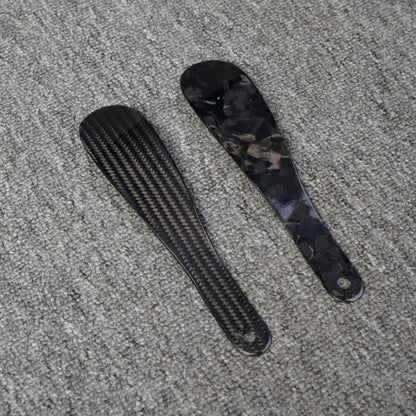 Close-up detail of two short carbon fiber shoe horns—one featuring a 3K carbon fiber woven pattern and the other showcasing a forged carbon fiber chopped texture—placed on a gray textured surface.