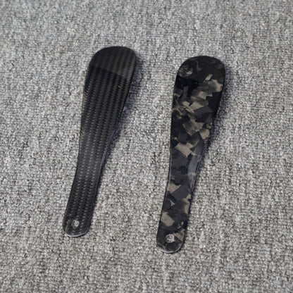 Close-up detail of two short carbon fiber shoe horns—one with a 3K carbon fiber woven pattern and the other with a forged carbon fiber chopped texture—placed on a gray textured surface.