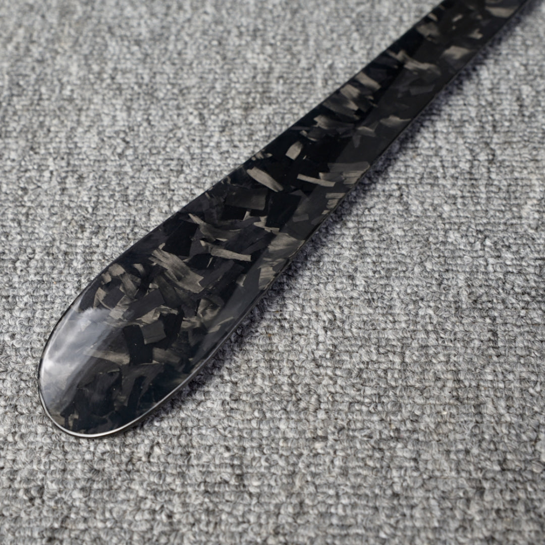 Close-up detail of a long forged carbon fiber shoe horn with a distinct chopped texture, placed on a gray textured surface.