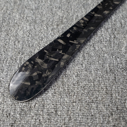 Close-up detail of a long forged carbon fiber shoe horn with a distinct chopped texture, placed on a gray textured surface.