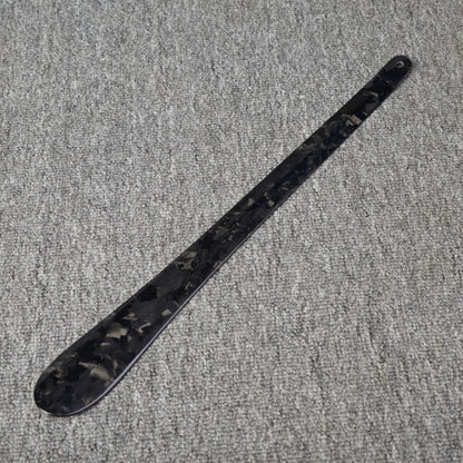 Close-up detail of a long forged carbon fiber shoe horn with a distinct chopped texture, placed on a gray textured surface.
