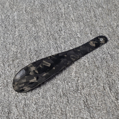 Close-up detail of a short forged carbon fiber shoe horn with a distinct chopped texture, placed on a gray textured surface.