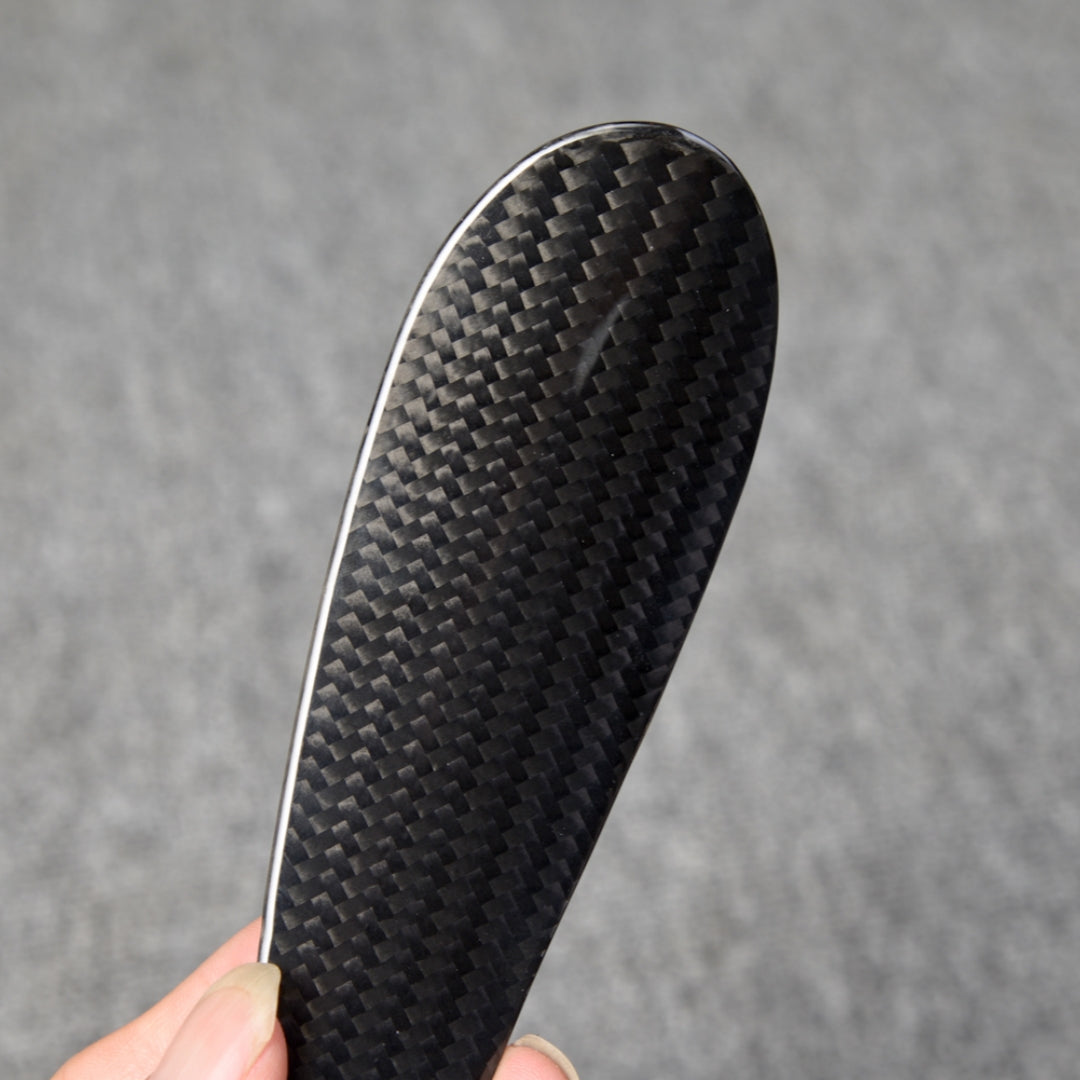 Close-up detail of a short 3K carbon fiber shoe horn with a distinct woven pattern, held in a hand against a gray textured background.