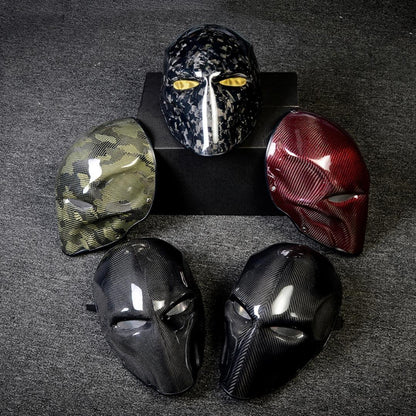 Close-up of multiple carbon fiber tactical masks with various designs — including camouflage pattern, glossy black, and red carbon fiber finishes — displayed on a dark textured surface.