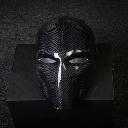 Close-up of a 3K twill carbon fiber tactical mask with mesh eye openings and a glossy finish, displayed on a black box against a dark textured background.