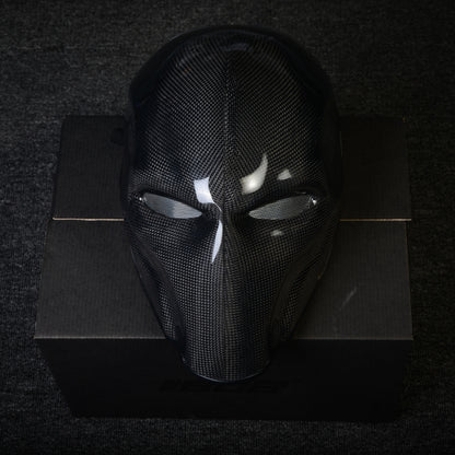 Close-up of a 3K plain weave carbon fiber tactical mask with mesh eye openings and a glossy finish, displayed on a black box against a dark textured background.