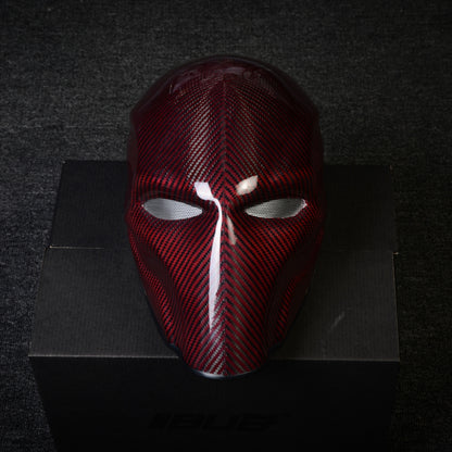 Close-up of a red 3K twill carbon fiber tactical mask with mesh eye openings and a glossy finish, displayed on a black box against a dark textured background.