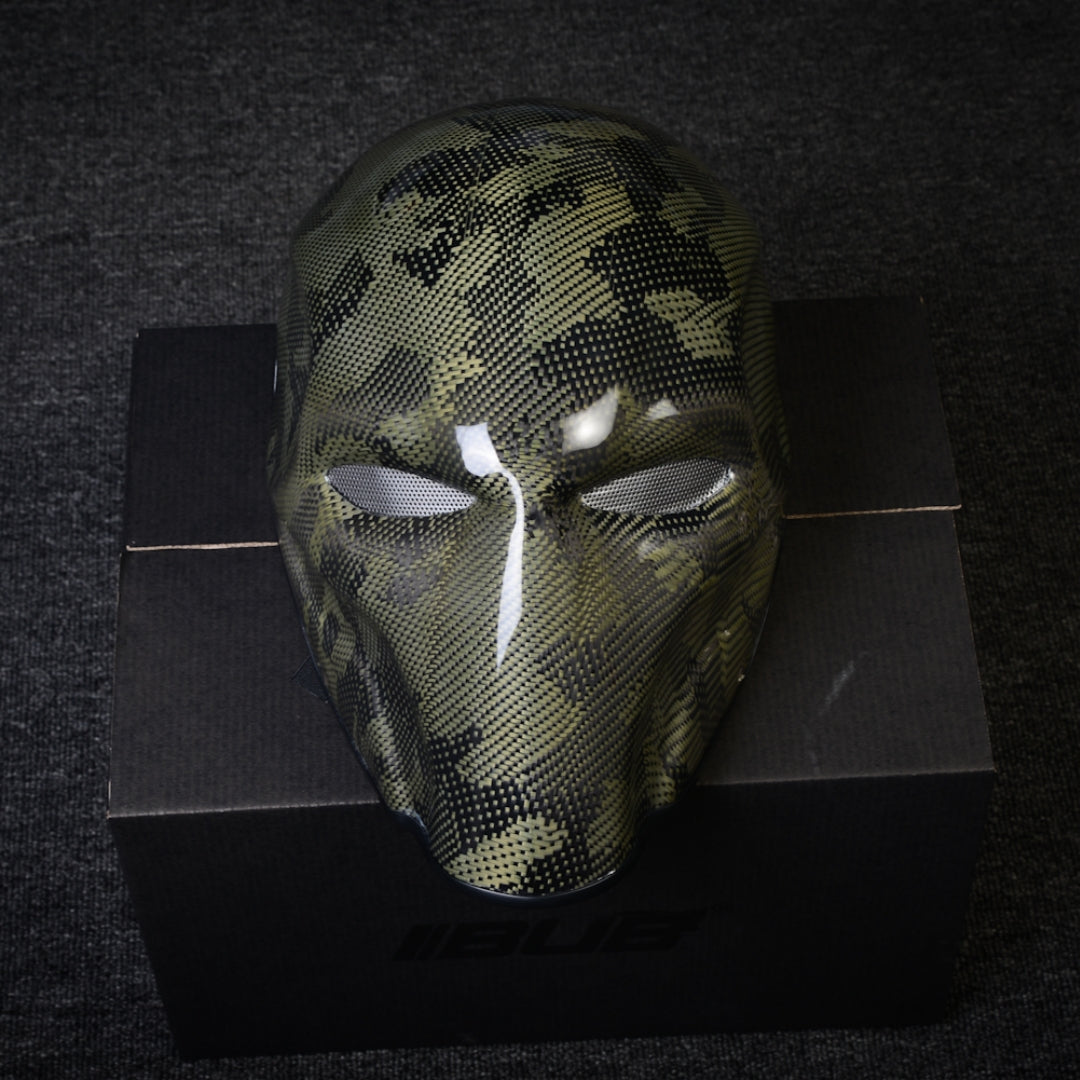Close-up of a forged carbon fiber tactical mask with a camouflage pattern, mesh eye openings, and a glossy finish, displayed on a black box against a dark textured background.