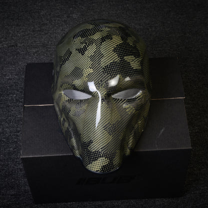 Close-up of a forged carbon fiber tactical mask with a camouflage pattern, mesh eye openings, and a glossy finish, displayed on a black box against a dark textured background.