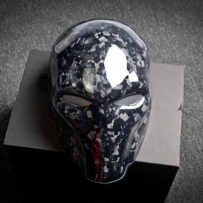Close-up of a forged carbon fiber tactical mask with a speckled black-and-white pattern, mesh eye openings, and a glossy finish, displayed on a dark box against a textured gray background.