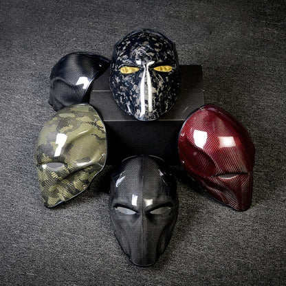 Close-up of multiple carbon fiber tactical masks in various designs — including camouflage, speckled, glossy black, and red carbon fiber patterns — arranged on a dark textured surface.
