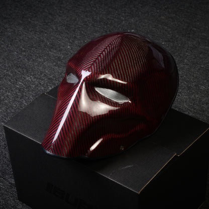 Close-up of a red 3K carbon fiber tactical mask with a glossy finish and mesh eye openings, displayed on a black box against a dark textured background.