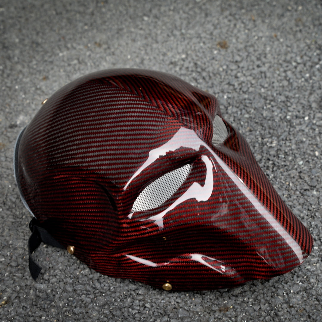 Close-up of a red 3K carbon fiber tactical mask with a glossy finish, featuring mesh eye openings, placed on a gravel surface.