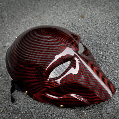Close-up of a red 3K carbon fiber tactical mask with a glossy finish, featuring mesh eye openings, placed on a gravel surface.