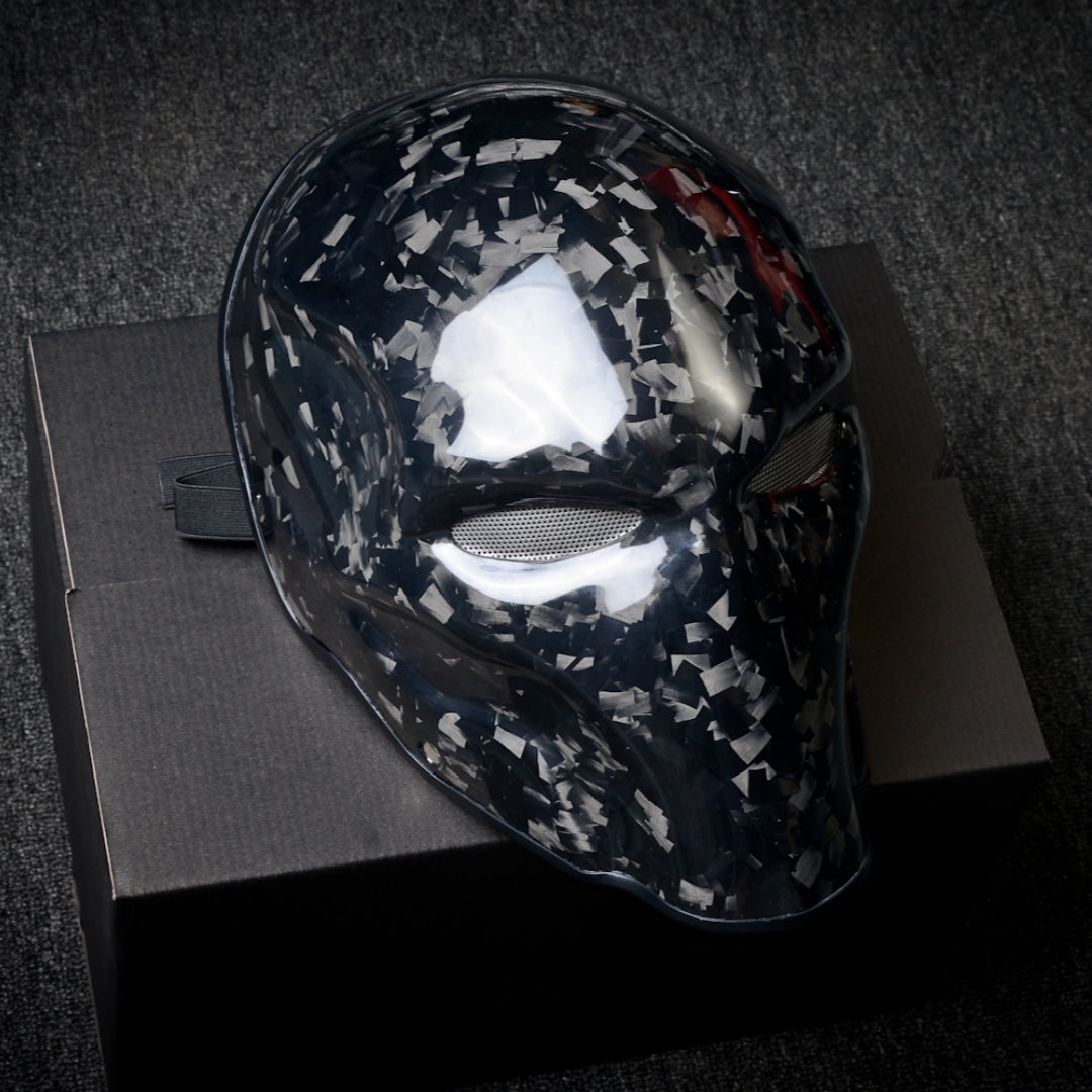 Close-up of a forged carbon fiber tactical mask with a speckled black-and-white pattern, mesh eye openings, and a glossy finish, displayed on a black box against a dark textured background.