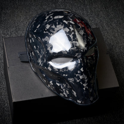 Close-up of a forged carbon fiber tactical mask with a speckled black-and-white pattern, mesh eye openings, and a glossy finish, displayed on a black box against a dark textured background.