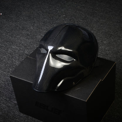 Close-up of a black 3K twill carbon fiber tactical mask with a glossy finish and mesh eye openings, displayed on a black box against a dark textured background.
