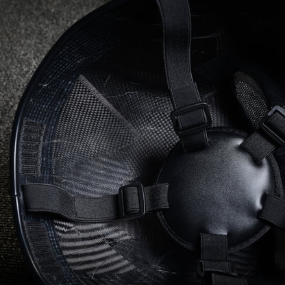 Close-up of the interior of a 3K twill carbon fiber tactical mask, showcasing black adjustable straps with buckle fasteners and the visible carbon fiber pattern through the inner surface, against a textured background.