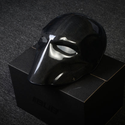 Close-up of a black 3K plain weave carbon fiber tactical mask with a glossy finish and mesh eye openings, displayed on a black box against a dark textured background.