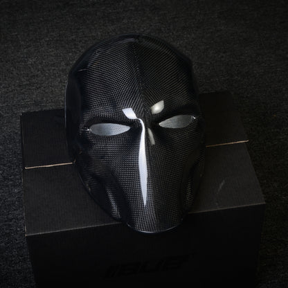 Close-up of a black 3K plain weave carbon fiber tactical mask with a glossy finish and mesh eye openings, displayed on a black box against a dark textured background.