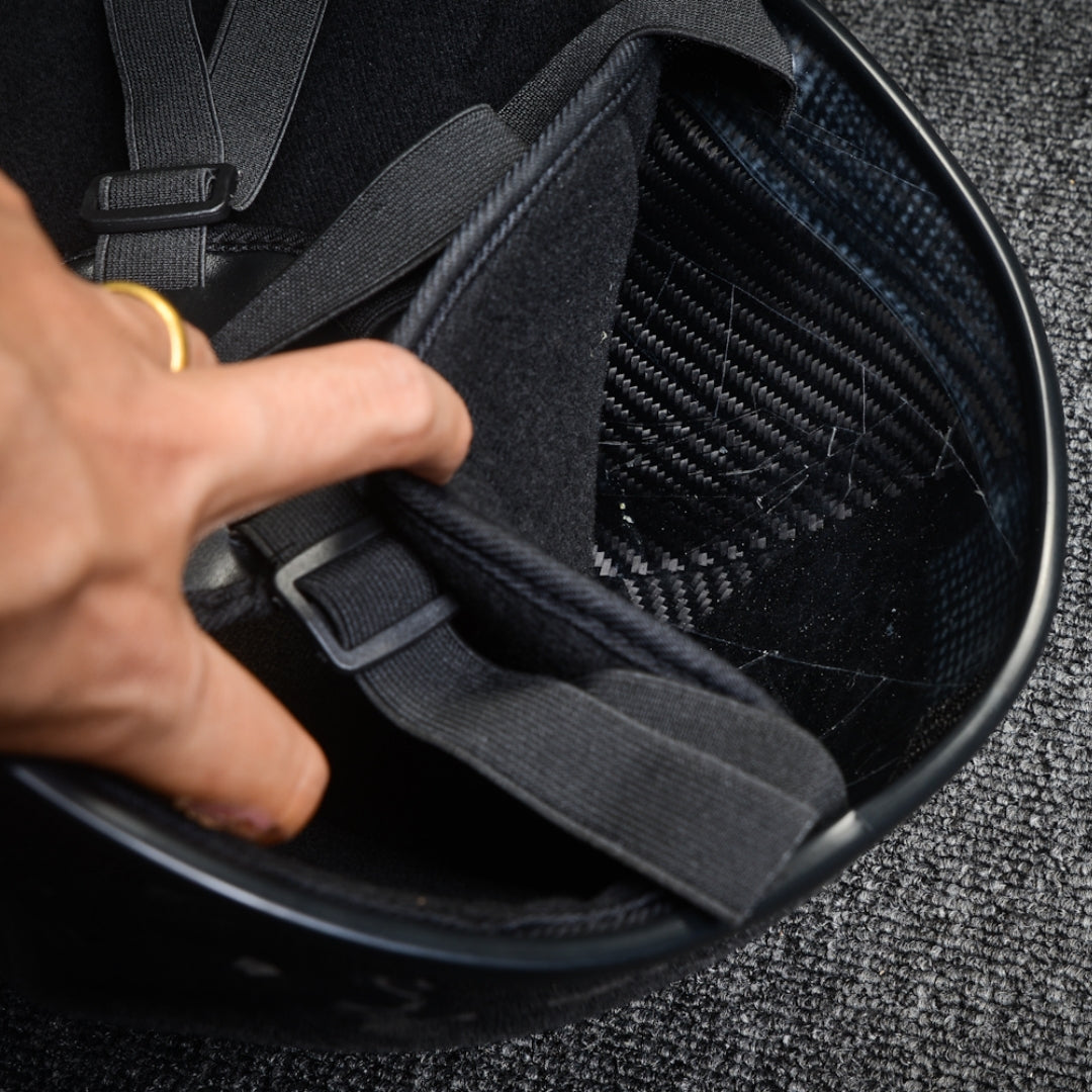 Close-up of the interior of a 3K plain weave carbon fiber tactical mask, showing black padding, adjustable straps with buckles, visible carbon fiber pattern, and a hand adjusting the components, against a textured background.