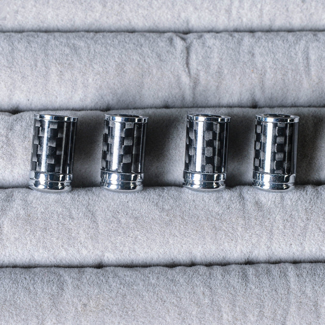 Close-up of four carbon fiber car tire valve stem caps with chrome detailing, arranged on a light gray textured fabric surface.