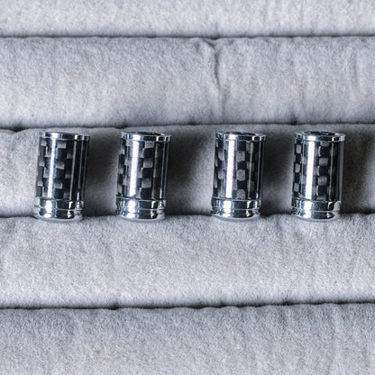 Close-up of four carbon fiber car tire valve stem caps with chrome detailing, arranged on a light gray textured fabric surface.