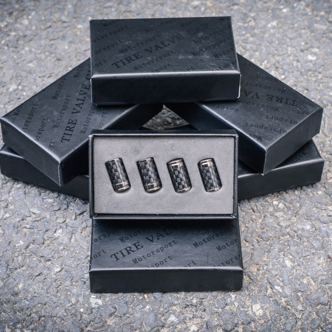 Close-up of four carbon fiber tire valve caps packaged in an open black box, with stacked boxes labeled "TIRE VALVE CAP" and "Motorsport" in the background, set on a rough asphalt surface.