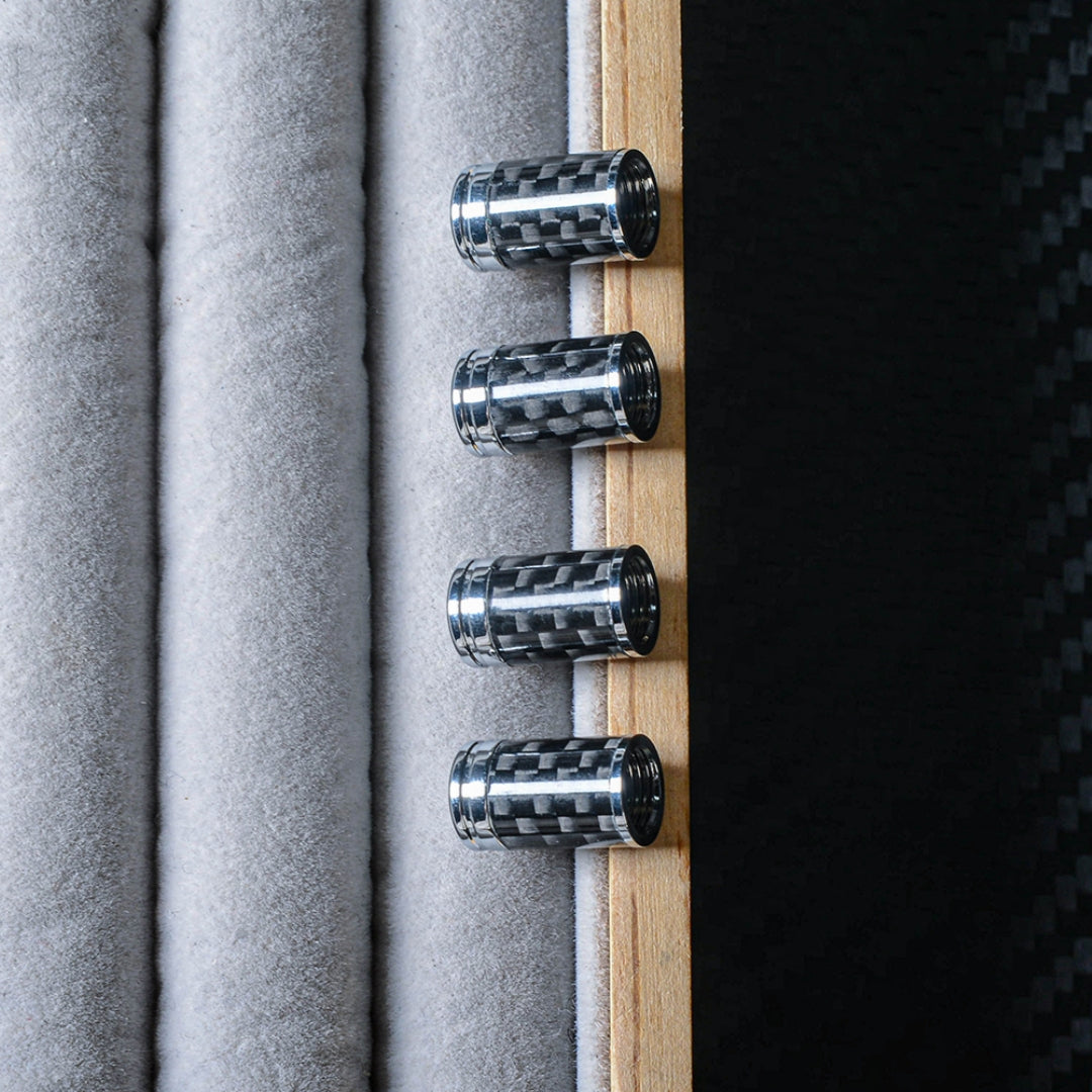 Close-up of four carbon fiber car tire valve stem caps with chrome accents, arranged vertically against a light gray velvet-like and wooden backdrop, with a carbon fiber textured surface visible on the right.