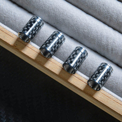Close-up of four carbon fiber car tire valve stem caps with chrome detailing, lined up on a wooden edge with gray velvet padding, showcasing the carbon fiber pattern against a dark carbon fiber-textured background.
