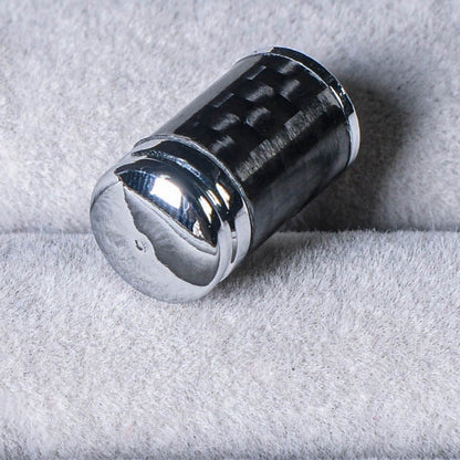 Close-up of a carbon fiber car tire valve stem cap with a shiny chrome-plated top, showcasing the carbon fiber pattern, resting on a soft gray textured surface.