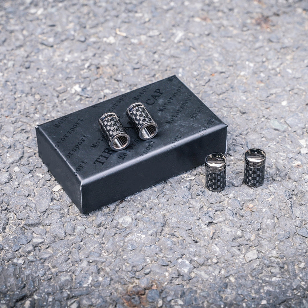 Close-up of four carbon fiber car tire valve stem caps with a checkered pattern, displayed on a black packaging box labeled "TIRE CAP" and "Motorsport," set against a rough asphalt surface.