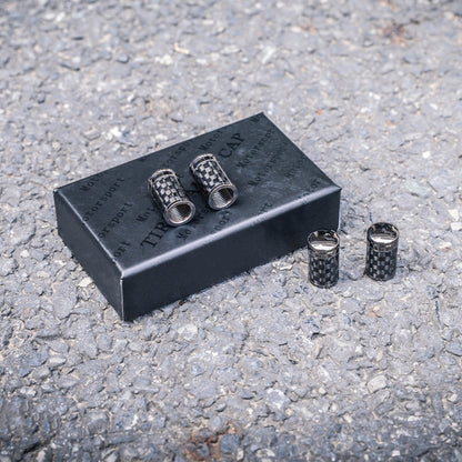 Close-up of four carbon fiber car tire valve stem caps with a checkered pattern, displayed on a black packaging box labeled "TIRE CAP" and "Motorsport," set against a rough asphalt surface.